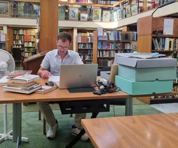 Someone sitting in the Library with books, laptop and archive boxes on the desk