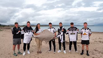 Seven people standing on a beach with the sea in the background holding a shark model and a football shirt