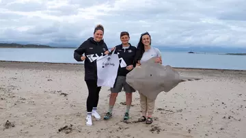 Three people standing on a beach with the sea in the background holding a shark model and a football shirt