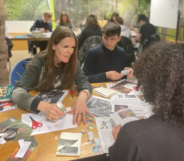 Linder Sterling sitting at a table with school children and scrap book cuttings
