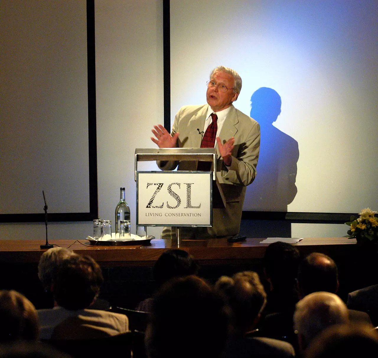 Sir David Attenborough standing at a ZSL branded podium delivering a lecture to an audience