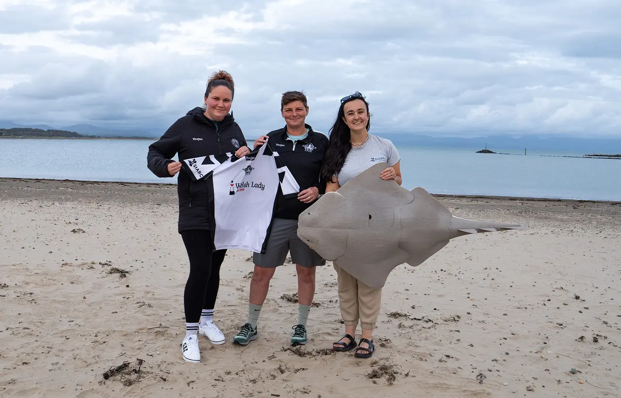 Three people standing on a beach with the sea in the background holding a shark model and a football shirt
