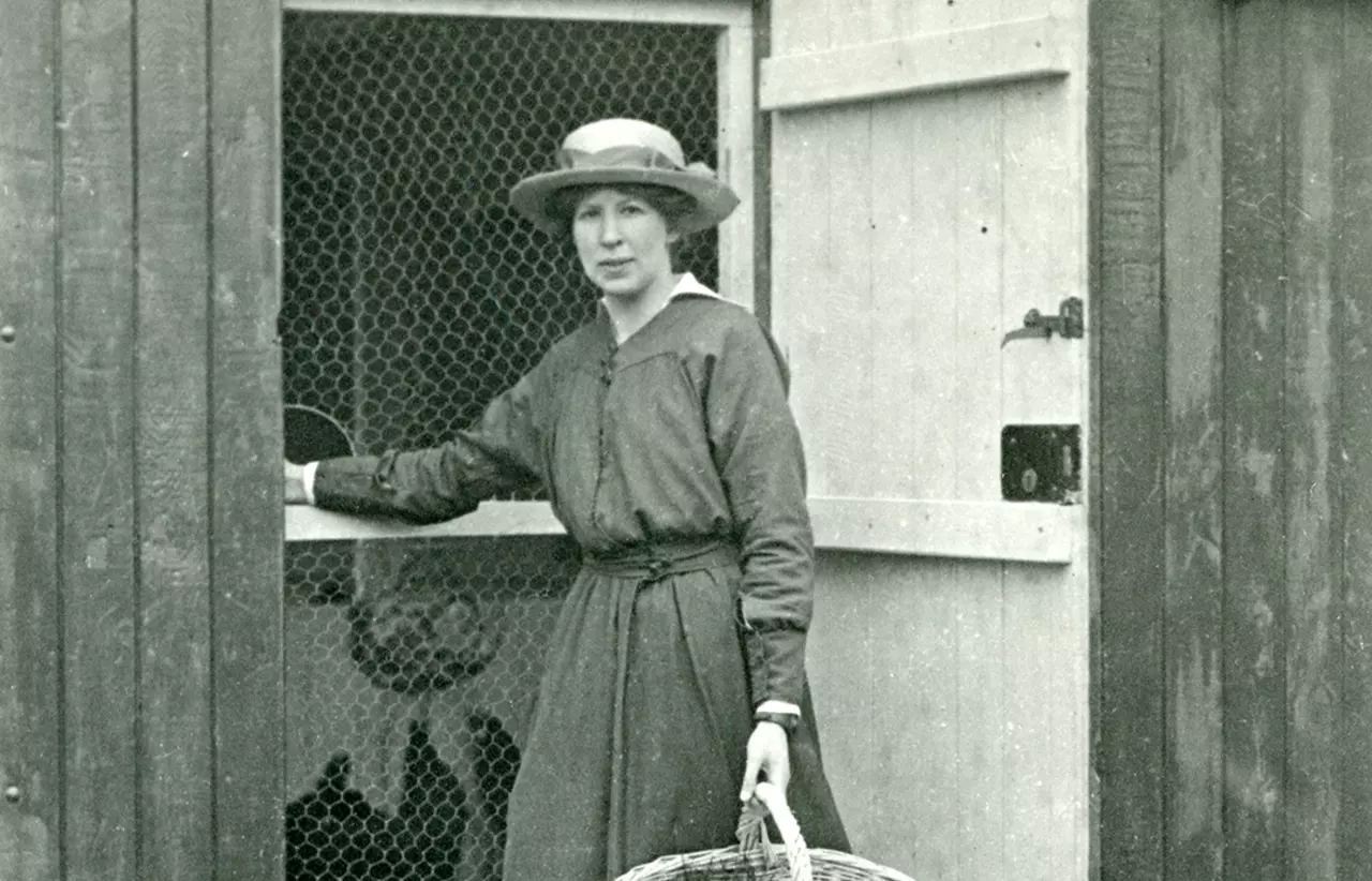A black and white photograph of Marion Saunders standing in front of a chicken shed holding a basket of eggs