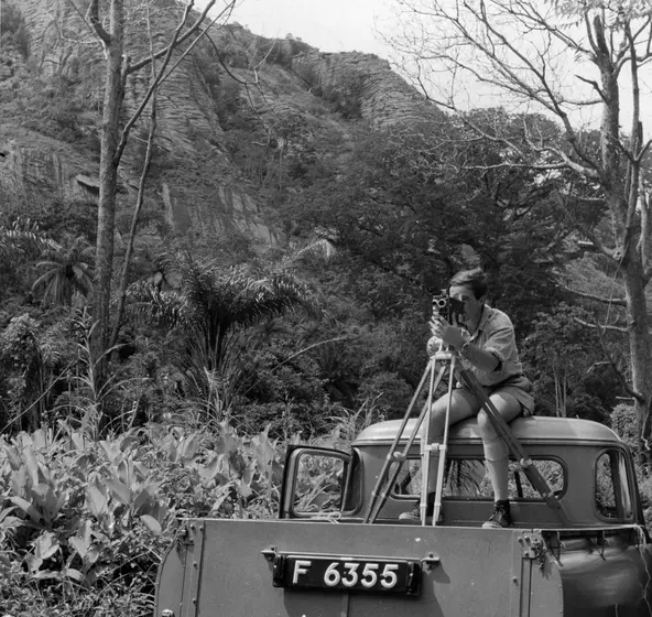 A black and white photograph of Charles Lagus sitting on the back of a truck filming Zoo Quest
