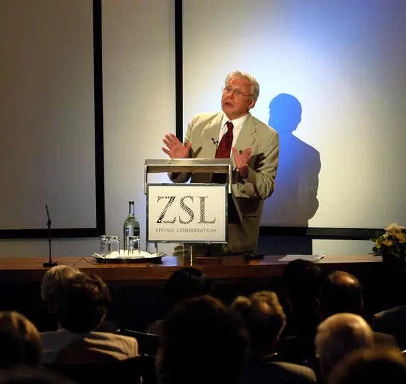 Sir David Attenborough standing at a ZSL branded podium delivering a lecture to an audience